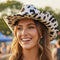 A smiling woman outdoors wearing a white cowboy hat with black cow-print spots, photographed in warm natural light with blurred trees and people in the background.