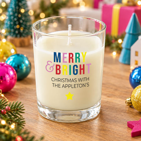 A white wax candle in a clear glass jar sits on a wooden table surrounded by Christmas decorations, including ornaments, lights, and wrapped gifts. The front of the candle reads 'MERRY & BRIGHT CHRISTMAS WITH THE APPLETON'S' with colorful lettering and a small yellow star.