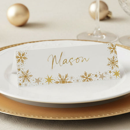 A white personalized place card with gold foil snowflakes and the name 'Mason' in gold script, displayed on a white plate with a gold charger and holiday ornaments in the background.