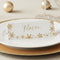 A white personalized place card with gold foil snowflakes and the name 'Mason' in gold script, displayed on a white plate with a gold charger and holiday ornaments in the background.