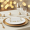 A festive table setting with a white place card featuring a gold snowflake border, layered over a gold charger plate on a beige tablecloth. Gold wine glasses, a gold fork, and decorative gold baubles are arranged in the background with warm bokeh lights.