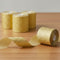 Gold textured ribbon rolls on a wooden surface, with one roll in the foreground showing a long strip of shiny metallic gold ribbon uncoiled.