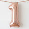 A metallic rose gold foil balloon shaped like the number '1', hanging from a matching rose gold ribbon against a plain white background.