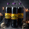Three large plastic bottles filled with very dark liquid, each with a purple cap and a yellow caution-style label reading 'CAUTION CONTAMINATED' and 'DRINK AT YOUR OWN RISK.' The bottles are arranged in a spooky Halloween setup with candles, cobwebs, skulls, and fog against a dark background.
