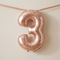 Air-filled rose gold number 3 balloon strung across a neutral wall with rose gold ribbon, soft light and shade, in square format