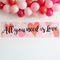 All you need is Love banner on a white wall with pink and red balloons cropped off the top