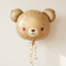 Bear balloon floating with gold curling ribbon against a cream wall with soft light and shade, in square format