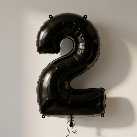 Black number 2 balloon styled to match the number 1, with black curling ribbon and soft light and shade against a neutral wall