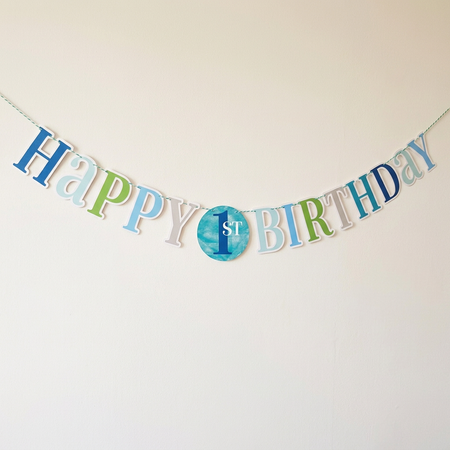 Blue and green first birthday bunting against an off-white wall with soft light and shade, in square format
