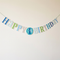 Blue and green first birthday bunting against an off-white wall with soft light and shade, in square format