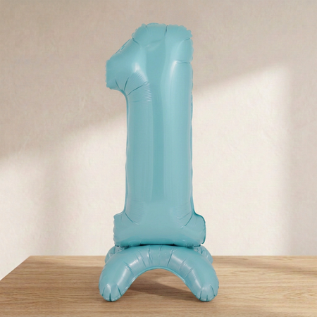 Blue number 1 standing balloon on a wooden tabletop with a cream background and soft light and shade, matching the pink balloon’s styling