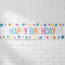 Colourful Happy Birthday star banner on a white brick wall with soft light and shade, in square format
