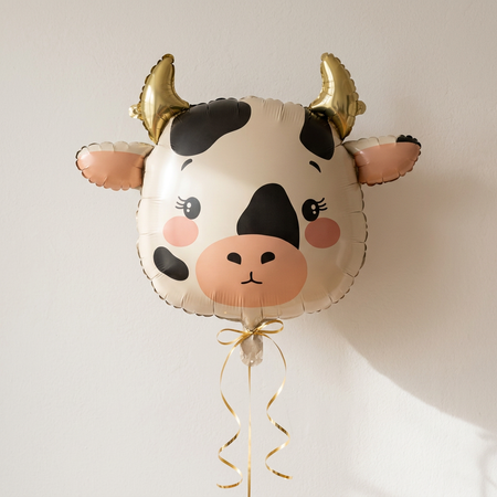Cow balloon floating with gold curling ribbon against a neutral wall with soft light and shade, close up in square format