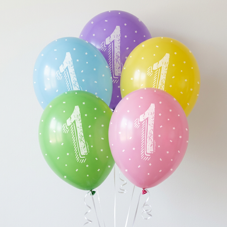 Five first birthday balloons floating with white curling ribbon against a softly lit white wall, in square format
