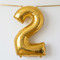 Gold number 2 balloon strung across a white wall with gold ribbon threaded through both top tabs, in square format with soft light and shade