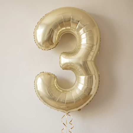 Gold number 3 balloon floating with gold curling ribbon against a softly lit neutral wall, in square format