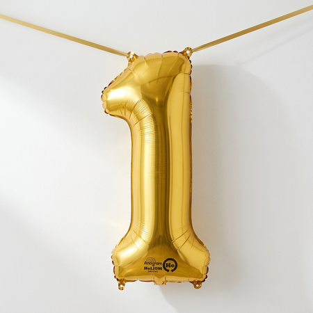 Gold number one balloon hung like bunting with gold ribbon threaded through its top holes against a softly lit white wall