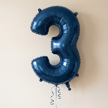 Navy number 3 balloon floating with navy curling ribbon against a softly lit neutral wall, in square format