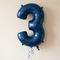 Navy number 3 balloon floating with navy curling ribbon against a softly lit neutral wall, in square format