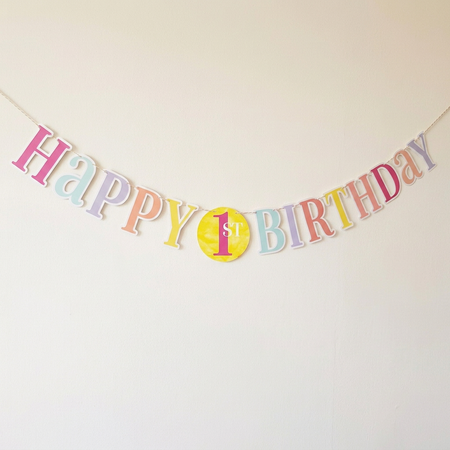 Pastel first birthday bunting against an off-white wall with soft light and shade, in square format