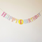 Pastel first birthday bunting against an off-white wall with soft light and shade, in square format