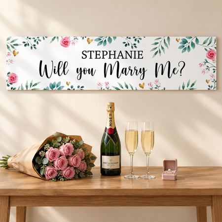 Proposal banner on a cream wall above a table with kraft-wrapped pink roses, champagne, two glasses and an engagement ring, proportioned to the 1.2m banner in square format