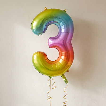 Rainbow number 3 foil balloon floating with gold curling ribbon against a neutral wall with soft light and shade