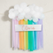 Rainbow streamer decoration and Olivia sign against an off-white wall with soft light and shade, in square format
