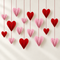 Red and pink paper hearts against an off-white wall with soft light and shade, with strings extending beyond the top of the square image