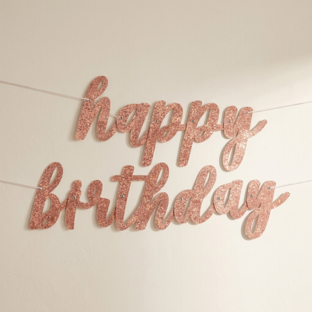 Rose gold happy birthday banner against an off-white wall with soft light and shade, in square format