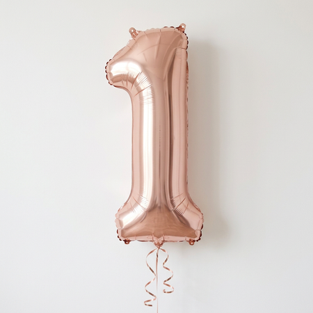 Rose gold number 1 balloon floating with rose gold curling ribbon against a white wall with soft light and shade, close up in square format