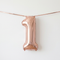 Rose gold number 1 balloon hanging from matching ribbon against a white wall with soft light and shade