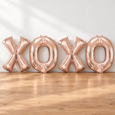 Rose-gold XOXO balloons resting on a wooden table against a white wall with soft light and shade, in square format
