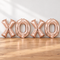 Rose-gold XOXO balloons resting on a wooden table against a white wall with soft light and shade, in square format