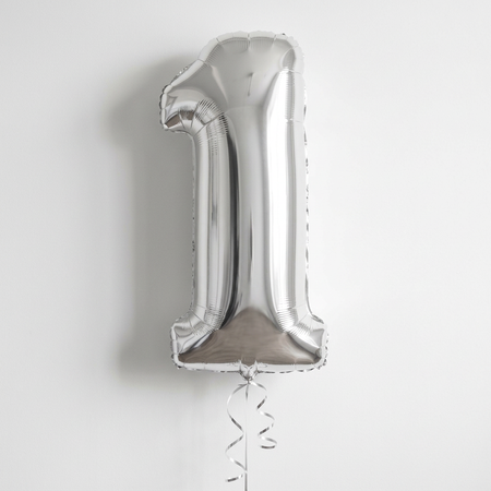 Silver number 1 balloon floating with silver curling ribbon against a softly lit white wall, close up in square format
