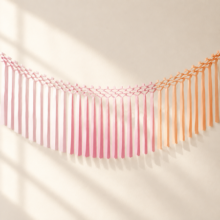 Square lifestyle image of the pink and orange fringe garland hanging across a softly lit wall